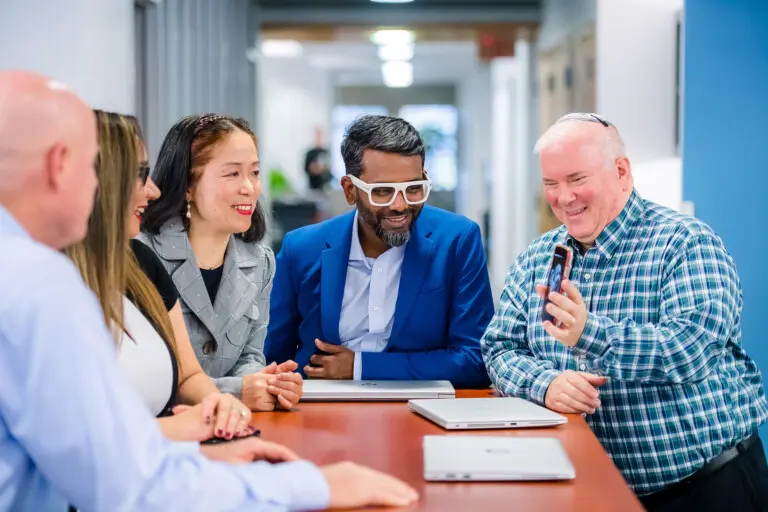 Five people in business casual attire sit around a small conference table and smile as they look at one man's phone screen.