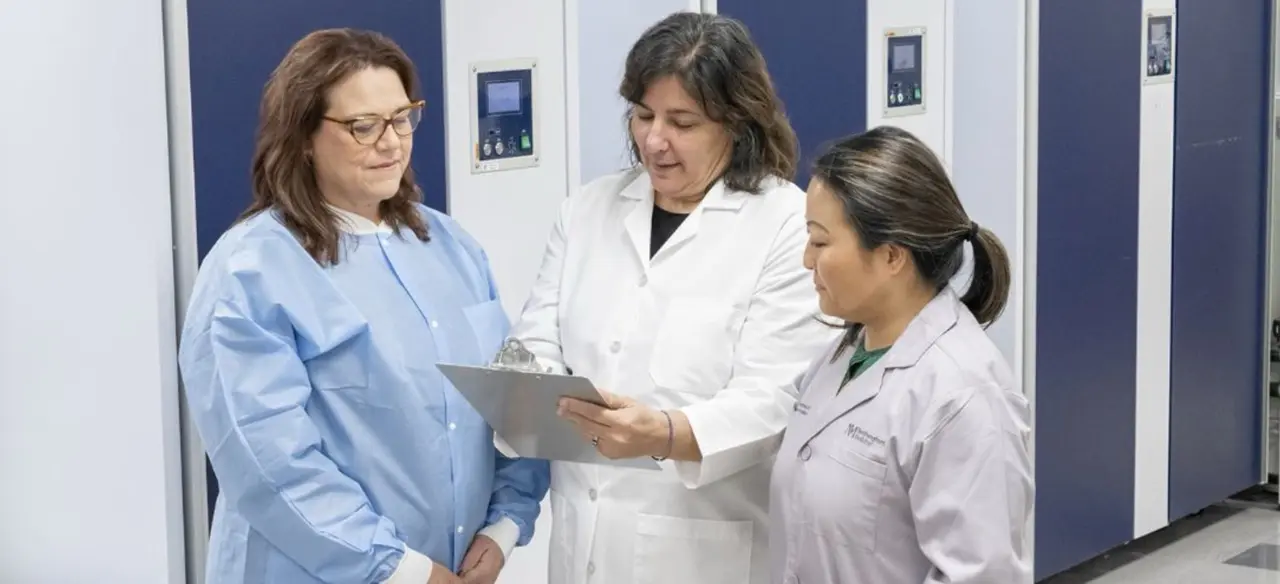 Two women in white lab coats and one woman in a blue medical gown look down toward a clipboard.