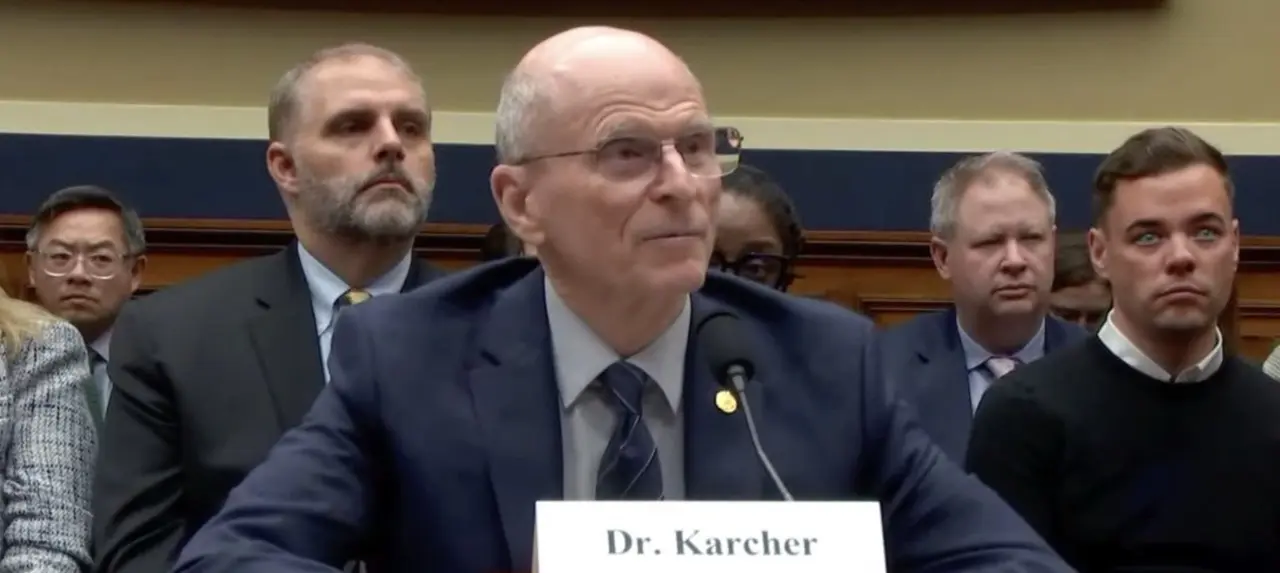 A man sits at a long desk with several people in business tire sitting behind him. In front of the man is a microphone and a name card that reads 'Dr. Karcher.'