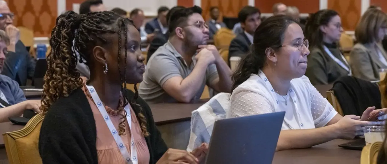Two women in professional wear and lanyards sit and watch a presentation in a room of over a dozen individuals.